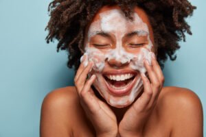 people, skin care and rejuvenation concept. pleased dark skinned woman with soft skin, applies white foam on face, touches cheeks, smiles broadly, cares of hygiene, feels relaxed and satisfied